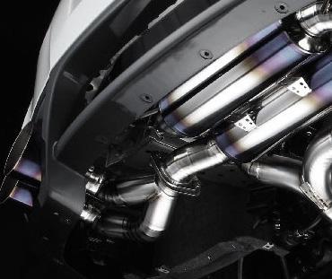Close-up view of a cars polished metal exhaust system showing multiple pipes and connectors. The metal has a reflective shine with areas of blue and purple discoloration, indicating heat exposure. The background is dark, accentuating the exhaust details.