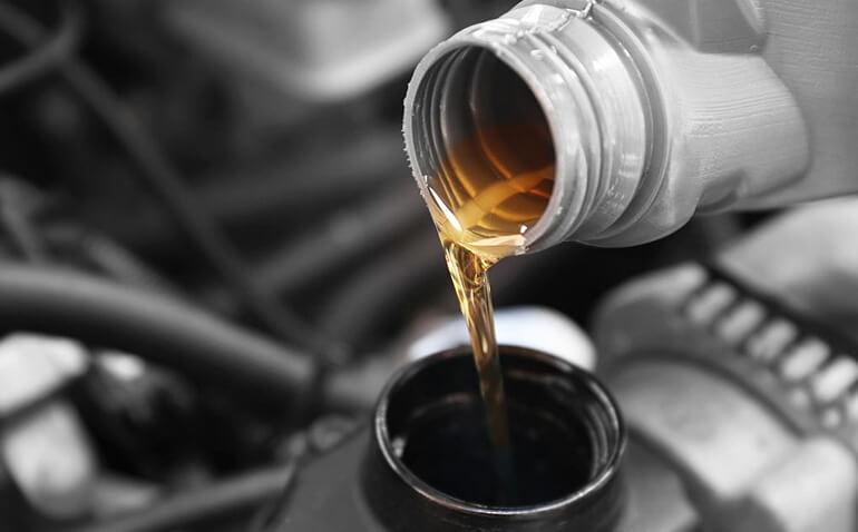 Close-up of motor oil being poured from a plastic container into a car engine, showing the oils golden color as it flows into the opening. The background is slightly blurred, highlighting the focus on the oil and the engine part.