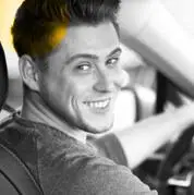 A person with short hair and a beard is smiling while sitting in the drivers seat of a car, looking over their shoulder. The image is mostly black and white with a yellow tint on their hair and a faint yellow background.