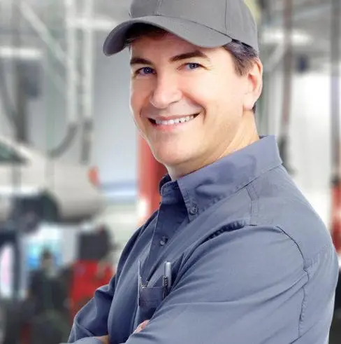 A smiling man in a gray cap and work uniform stands with arms crossed in a garage. Blurred background features cars and garage equipment.