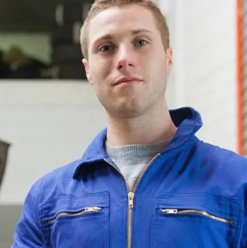 A person wearing a blue jacket with zippered pockets stands indoors, facing the camera. The background is slightly blurred, showing a mix of light and dark areas.