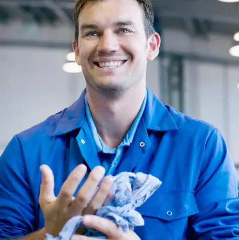 Smiling man in a blue work uniform stands inside an industrial setting, cleaning his hands with a cloth. The background is slightly blurred, highlighting the warehouse environment.