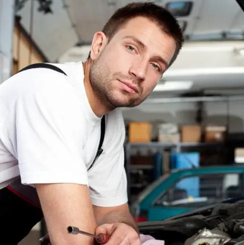 A man in a white t-shirt and overalls leans over a car engine in a garage. He holds a tool in his hand, looking towards the camera. The background shows shelves and another vehicle.