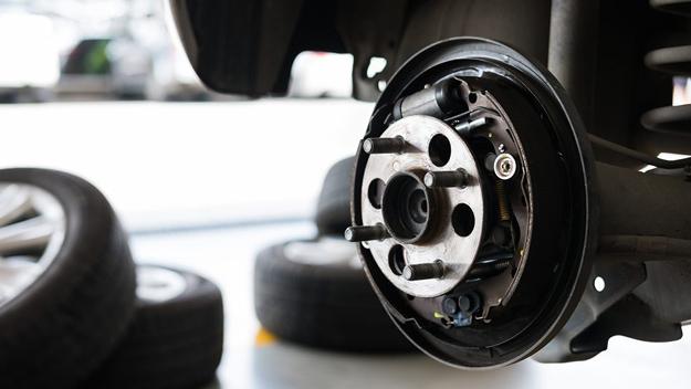 The Mufflerman, Mechanic Near Me Close-up of a cars rear drum brake assembly with the wheel removed, showcasing components like springs and wheel cylinder. Blurred tires are visible on the garage floor, hinting at ongoing maintenance such as wheel bearing replacement.