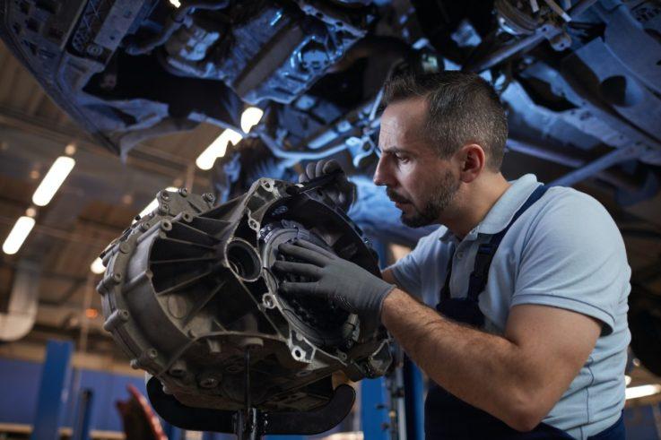 The Mufflerman A mechanic wearing gloves and overalls inspects a car part in a workshop. He is focused and examining the component closely, with an underside of a vehicle visible above him. The workshop is well-lit with industrial lighting.
