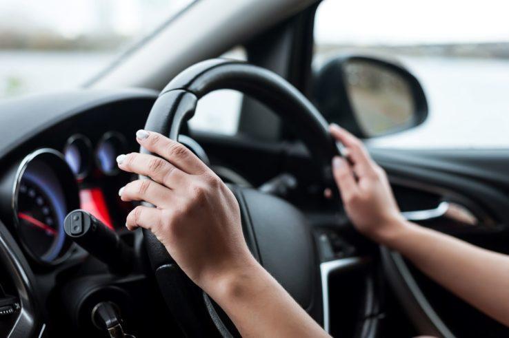 The Mufflerman, Mechanic Near Me Close-up of a person gripping a car steering wheel with both hands, sitting in the drivers seat. The dashboard showcases multiple dials, while the background reveals a blurred outdoor view through the windshield—experience precise alignment and steering with The Mufflerman expertise.