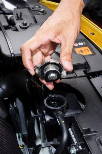 The Mufflerman, Mechanic Near Me A hand is holding a radiator cap above an open radiator in the vehicles engine cooling system. The cap is being removed or replaced, revealing the opening beneath it.