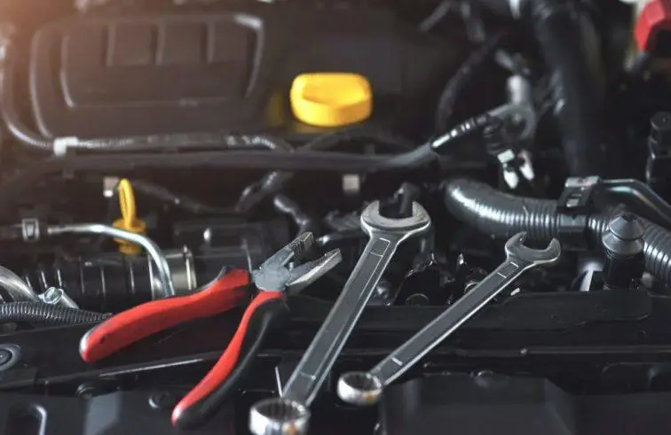 A close-up of an open car engine with a pair of pliers and two wrenches resting on it. The engine components are visible, and theres a yellow cap towards the back, surrounded by various tubes and wires.