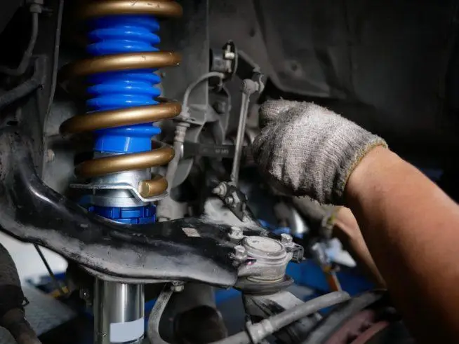 The Mufflerman, Mechanic Near Me A mechanics gloved hand expertly works on a cars suspension system, focusing intently on a blue and gold coilover spring, highlighting the precision of high-quality suspension services. The background reveals an array of mechanical components under the vehicle.