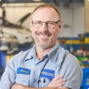 A mechanic in Stratford with glasses and a goatee stands smiling, arms crossed, in an auto repair shop. He wears a light blue shirt with a name tag reading Andy, against a backdrop of blurred machinery and tools.