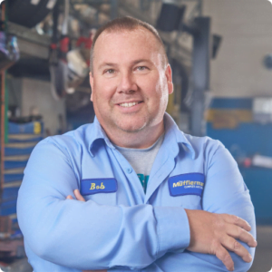 The Mufflerman, Mechanic Near Me A man in a blue uniform shirt with a name tag that reads Bob stands confidently with arms crossed in the bustling workshop of Cambridge Auto Shop. The background showcases shelves filled with tools and equipment. He is smiling warmly, embodying the local expertise youd expect from a mechanic near you.