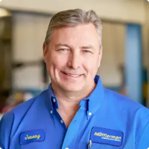 The Mufflerman A man with gray hair smiles at the camera, donning a blue work shirt with name tags. He stands in a Chatham Auto Repair setting, surrounded by blurred equipment, embodying the friendly expertise of your go-to mechanic in Chatham.