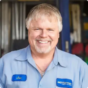 The Mufflerman A smiling man with light hair and a beard stands in a workshop. Clad in a light blue uniform with a name tag reading Joe and a logo patch, he embodies the trusted auto repair specialist in St. Thomas, ready to assist you nearby amidst the blurred backdrop of tools and equipment.