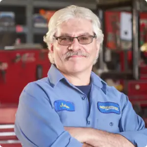 The Mufflerman A person with gray hair, a mustache, and glasses smiles confidently, standing with arms crossed. They wear a blue work shirt adorned with John and a company name on patches. The backdrop reveals an auto repair workshop in London South, complete with red tool cabinets.