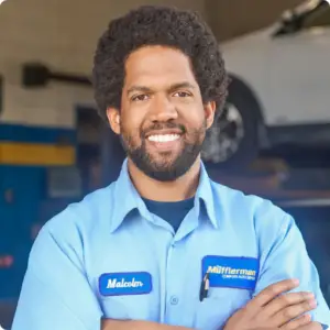 The Mufflerman In an auto repair shop, a mechanic in Stratford wearing a light blue work shirt smiles. His patch reads Malcolm and Mufflerman, with a blurred car lifted in the background.