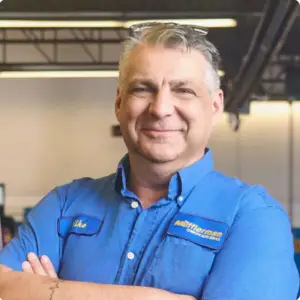 The Mufflerman, Mechanic Near Me A smiling person in a blue collared shirt with embroidered patches stands indoors, embodying the heart of Brantford Auto Repair. The garage backdrop, with its visible ceiling beams and lighting, hints at their expertise as a trusted mechanic near me.