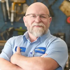 A bearded man with glasses, wearing a light blue uniform with a name tag, stands confidently with arms crossed. The background showcases blurred tools on a pegboard, hinting at his expertise in Kitchener Auto Repair.