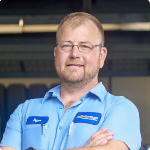 The Mufflerman A mechanic with short hair and glasses is smiling. He wears a light blue shirt with embroidered name tags, hinting at his role in auto repair in Stratford, and has a pen in his pocket. The background is a blurred indoor setting.