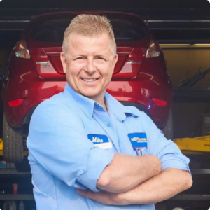 The Mufflerman, Mechanic Near Me A smiling person in a blue mechanic shirt stands confidently with arms crossed in front of a red car elevated on a lift inside an auto repair shop. The name tag reads Mike, your trusted mechanic near me in Sarnia for all auto repair needs.