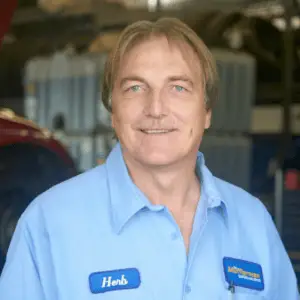 The Mufflerman, Mechanic Near Me Middle-aged man with light brown hair and a mustache, wearing a blue button-up shirt with a name tag reading Herb and a Mufflerman patch. He is smiling in an automotive repair workshop, suggesting hes the go-to mechanic near me in Sarnia for reliable auto repair services.