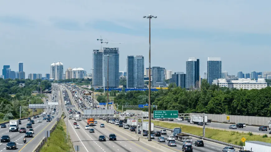 The Mufflerman, Mechanic Near Me A busy multi-lane highway with numerous cars and trucks flows toward a distant skyline of tall buildings under a clear blue sky. Green trees line the road, newly restored after pothole damage repair in Southern Ontario. Overhead signs provide directions as cranes rise among the skyscrapers.
