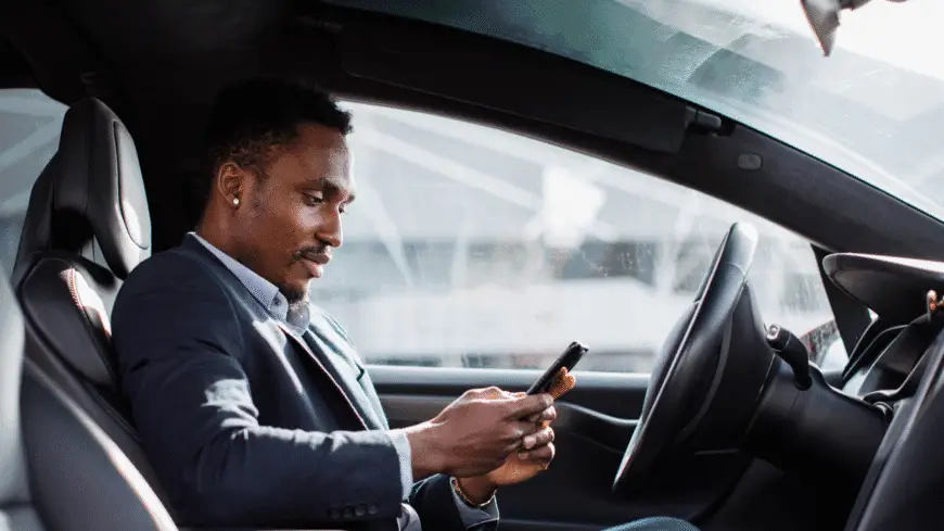 The Mufflerman, Mechanic Near Me A man in a suit sits in the driver’s seat of a car, looking at and using his smartphone, with daylight shining through the windows.
