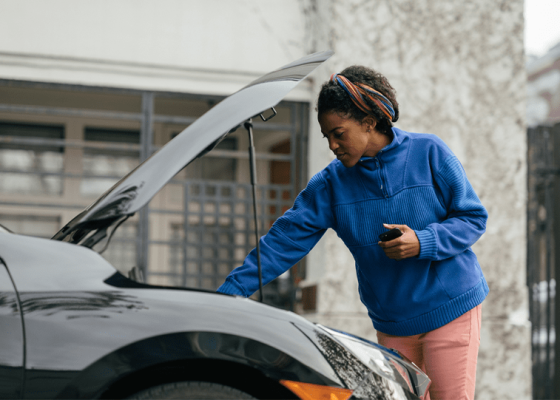 The Mufflerman A woman in a blue sweater and pink pants inspects under the open hood of a black car parked on the street. With her phone in hand, she seems concerned about one of those 6 vehicle noises you shouldnt ignore. In the background, a building with a barred window stands quietly.