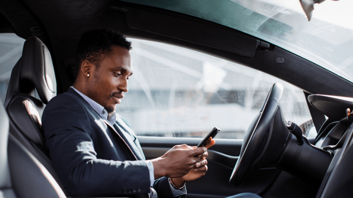 The Mufflerman, Mechanic Near Me A man in a suit sits in the driver’s seat of a car, looking at and using his smartphone, with daylight shining through the windows.