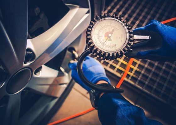 The Mufflerman Person in blue gloves checks car tire pressure with a gauge attached to an air hose, emphasizing one of the 7 Important Spring Car Maintenance Tips. The gauge is close to the camera, showing pressure dials, while the tire and wheel rim are visible on the left.