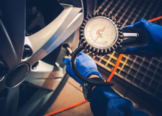 The Mufflerman Person in blue gloves checks car tire pressure with a gauge attached to an air hose, emphasizing one of the 7 Important Spring Car Maintenance Tips. The gauge is close to the camera, showing pressure dials, while the tire and wheel rim are visible on the left.
