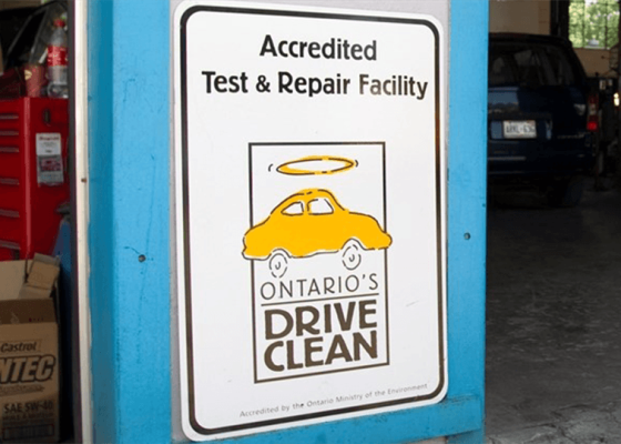 The Mufflerman, Mechanic Near Me Sign for Ontarios Drive Clean, an accredited test and repair facility, displayed on a wall. As Drive Clean is ending, and heres what you need to know: the sign features a yellow car with a halo above it against a backdrop of garage parts and a partially visible car.