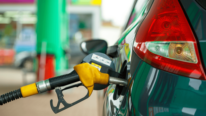 The Mufflerman, Mechanic Near Me A close-up of a yellow gasoline pump nozzle inserted into the fuel tank of a dark green car at a gas station. The car’s taillight is visible, and the background shows a blurred view of the station.