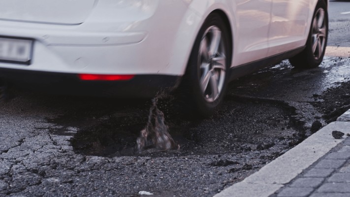 The Mufflerman, Mechanic Near Me A white car drives through a large pothole, splashing water onto the road in Southern Ontario. The pothole, marked for upcoming repair, is surrounded by cracked asphalt along the edge of a sidewalk.