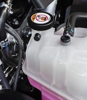 The Mufflerman Close-up of a car engine compartment highlighting the radiator and engine cooling system service components. A coolant reservoir with a black cap and warning label holds pink fluid, surrounded by various hoses and metal components.