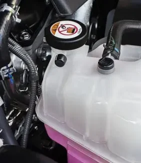 The Mufflerman Close-up of a car engine compartment highlighting the radiator and engine cooling system service components. A coolant reservoir with a black cap and warning label holds pink fluid, surrounded by various hoses and metal components.