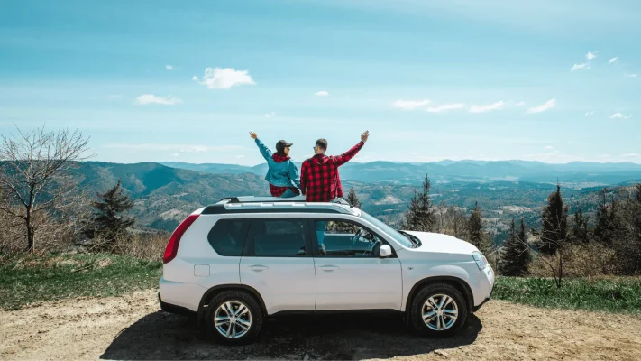 The Mufflerman, Mechanic Near Me Two people sit on the roof of a parked white SUV, arms lifted in excitement. They overlook a scenic view of rolling mountains and a clear blue sky, savoring the sunny day—a brief escape before addressing lifes little hurdles like pothole damage repair in Southern Ontario.