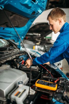The Mufflerman, Mechanic Near Me The mechanic in a blue uniform diligently works under the hood, using precise tools on the battery. A digital multimeter lies nearby as he performs a car battery repair. The setting is a bustling garage, showcasing an expert car battery service near me.