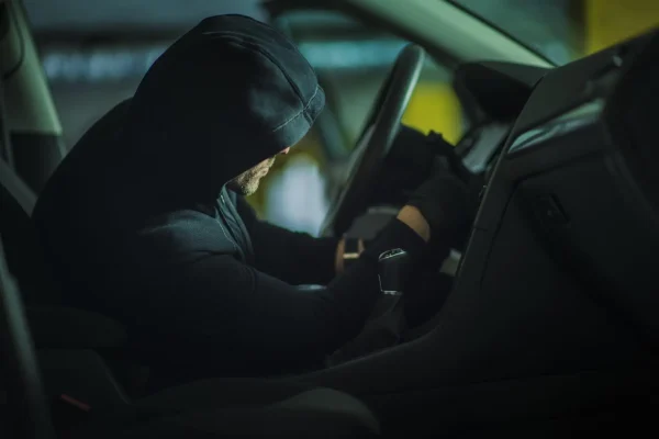 The Mufflerman, Mechanic Near Me A person in a black hoodie and gloves sits in the drivers seat of a car, appearing to tamper with the ignition. The dimly lit scene underscores the importance of knowing how to prevent auto theft, as secrecy and stealth loom large.