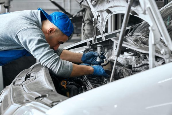 The Mufflerman In the workshop, a mechanic in a blue cap and gloves carefully inspects the car engine under the open hood, looking for any of the 10 most common engine light issues.