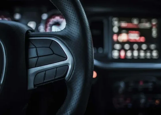 The Mufflerman Close-up of a car’s steering wheel with buttons visible, hinting at important safety tips about driver assistance technologies. In the background, a blurry dashboard with illuminated digital displays and controls. The dimly lit interior exudes a modern, high-tech feel.