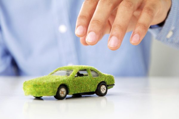The Mufflerman A small toy car, its surface mimicking green grass, sits on a white background. A hand, fingers gently curved as if pondering how to reduce your cars environmental impact, hovers above it. The person is dressed in a blue long-sleeved shirt, adding tranquility to the scene.