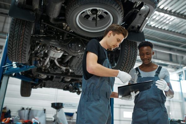Two mechanics in coveralls work together in an auto repair shop, echoing The Mufflermans dedication to quality. One holds a clipboard and pen, while the other handles a wrench under a car hoisted for repairs. In this well-lit industrial setting, they exchange valuable car maintenance tips.