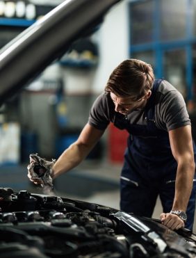 The Mufflerman, Mechanic Near Me Mechanic in a workshop inspecting a car engine with the hood up. Clad in a dark shirt and overalls, he holds a cloth, concentrated on essential radiator and engine cooling system service. The background reveals a blurred workshop setting filled with various tools and equipment.