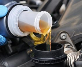 A person wearing blue gloves pours engine oil from a bottle into a car engine. The oil is amber-colored and flows smoothly into the opening. Visible parts of the engine surround the area where the oil is being poured.