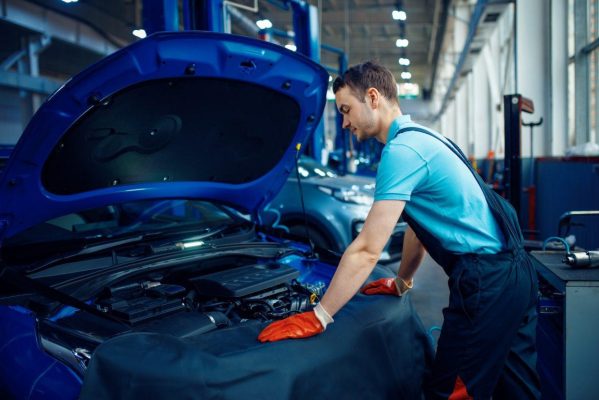 In a well-lit garage, a mechanic in a blue shirt and overalls carefully inspects the engine of a blue car. Sporting orange gloves, he stands next to various tools at The Mufflerman, ensuring every detail from alignment & steering is expertly handled.