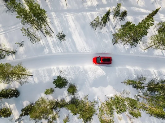 The Mufflerman, Mechanic Near Me A red car with winter tires drives along a snow-covered road surrounded by scattered evergreen trees, casting long shadows. The scene is viewed from above, highlighting the winding path through the winter landscape, as tire use rises in Ontario.