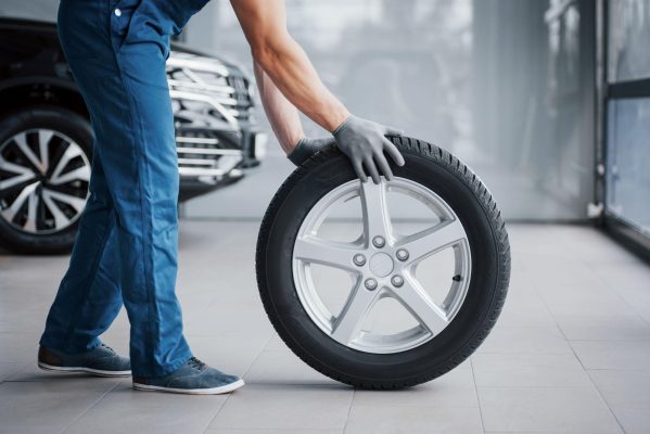 The Mufflerman, Mechanic Near Me In a bustling auto workshop, a person in blue coveralls and gloves expertly maneuvers a car tire upright. The backdrop features part of a car and gleaming glass surfaces, reminiscent of the comprehensive Summer Tires Guide—everything you need to know for choosing the best fit this season.