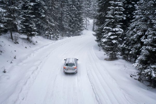 The Mufflerman, Mechanic Near Me A car is navigating a snow-covered road winding through a dense, snowy forest. Tall evergreen trees coated with snow line both sides of the road, creating a wintery scene that reminds one of the essential winter car maintenance tips needed under a gray, cloudy sky.