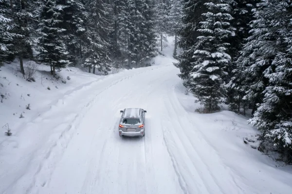 The Mufflerman, Mechanic Near Me A car is navigating a snow-covered road winding through a dense, snowy forest. Tall evergreen trees coated with snow line both sides of the road, creating a wintery scene that reminds one of the essential winter car maintenance tips needed under a gray, cloudy sky.