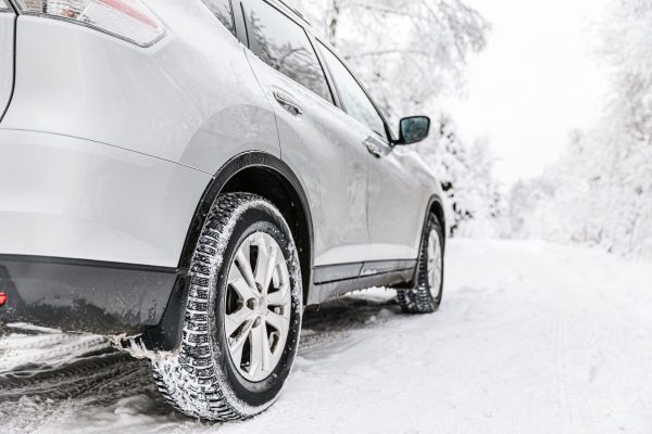 The Mufflerman, Mechanic Near Me A silver SUV with winter tire swaps is parked on a snowy road, surrounded by snow-covered trees. The focus is on the rear tire, highlighting its winter readiness, even during challenging times like COVID-19. The sky and surroundings are white and overcast.